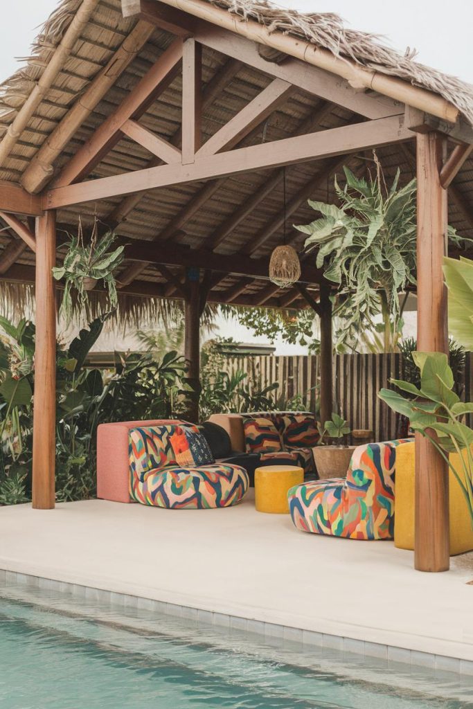 A tropical-style outdoor lounge with colorful chairs under a thatched gazebo surrounded by lush greenery near a pool.