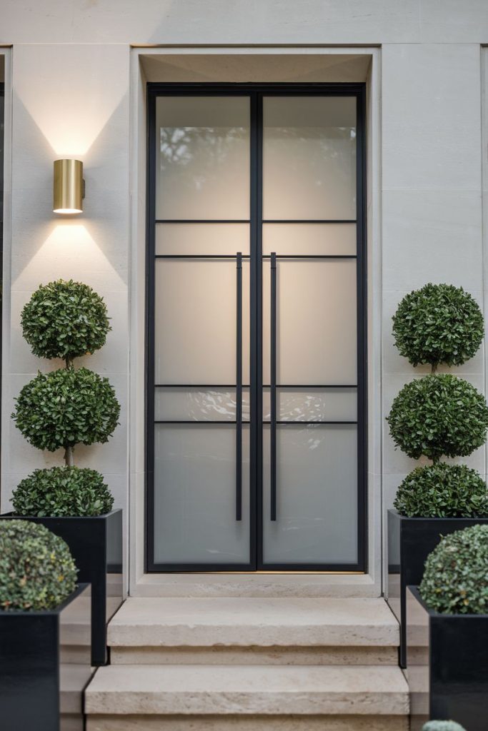 A modern glass door with a black frame and handles flanked by symmetrical topiary potted plants on stone steps, beneath a sconce.