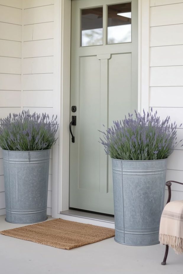 A white door with a window and a black handle is flanked by two metal planters filled with lavender. There is a wall lamp at the top and a bench with a checkered blanket on the side. There is a doormat in the front.