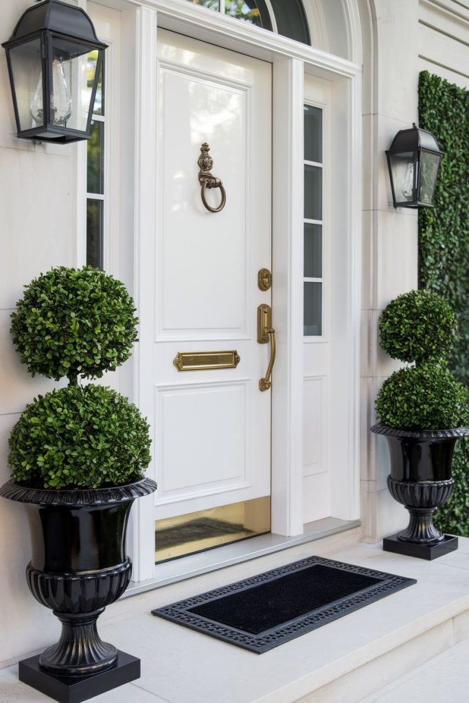A white front door with a brass knocker and mail slot flanked by large black planters with manicured topiary shrubs. There are two wall lanterns on each side of the door.