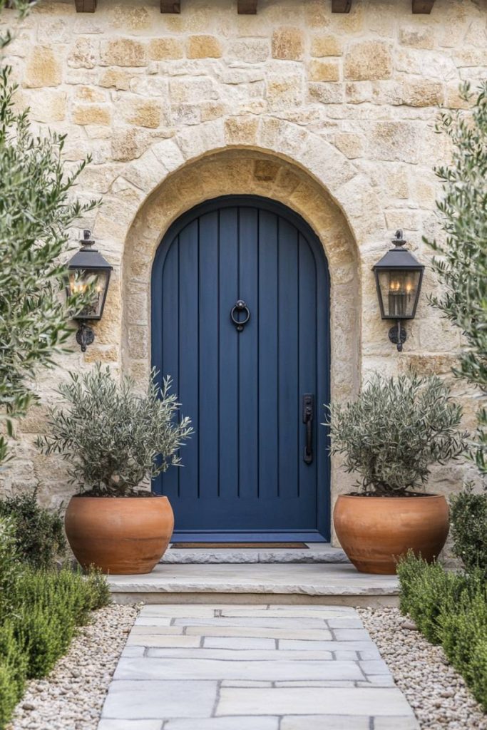 Blue arched wooden door in a stone wall flanked by two wall lanterns and large potted plants. A stone path leads to the entrance.