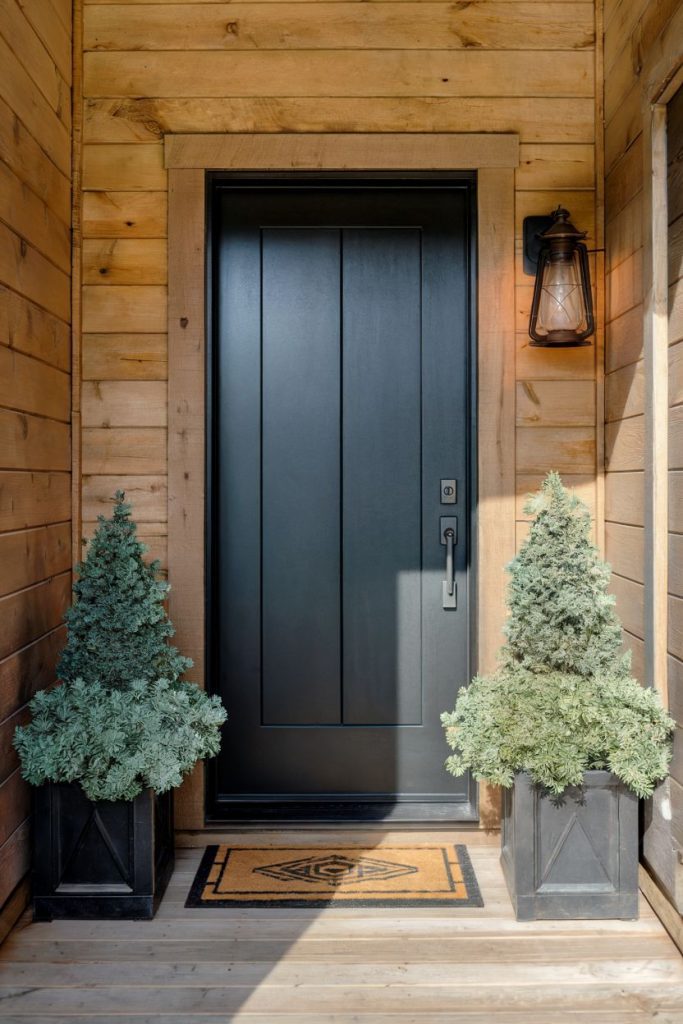 A black front door with a wooden frame flanked by two evergreen potted plants. A lantern light is mounted on the right. There is a welcome mat in front of it.