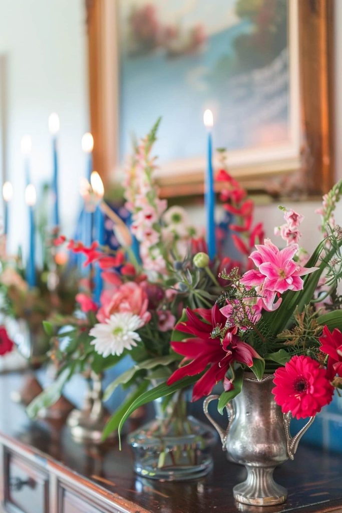 A close -up of a decorative arrangement with living flowers, including red, pink and white flowers, in silver vases. Blue taper candles are illuminated in the background and a framed painting is visible on the wall.