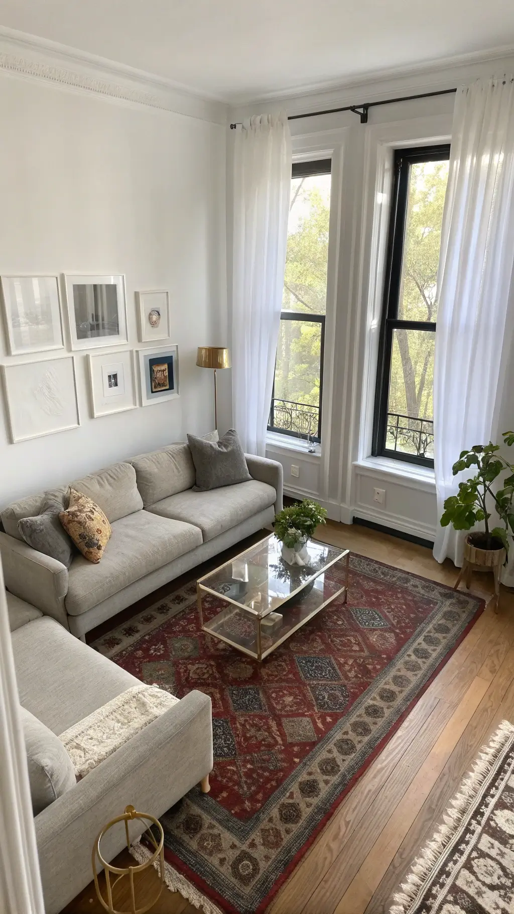 Overhead view of a sunlit 10x12' corner living room with a gray linen sofa, lucite nesting tables, white walls contrasted by black-framed windows, brass sculptures, vintage Persian rug, earthenware collection, and mirrors enhancing the sense of space.