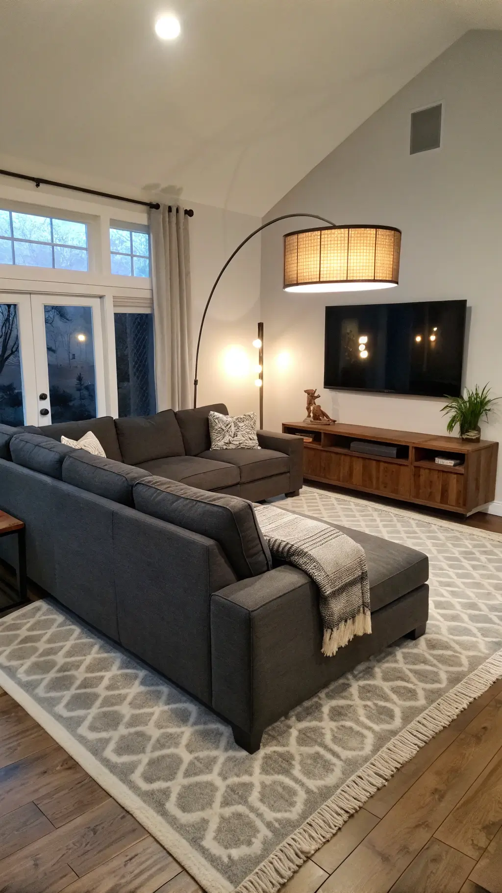 Modern living room with charcoal sectional sofa, arched floor lamp, wall-mounted sconces, walnut media console, and layered ivory and gray rugs, captured during blue hour.