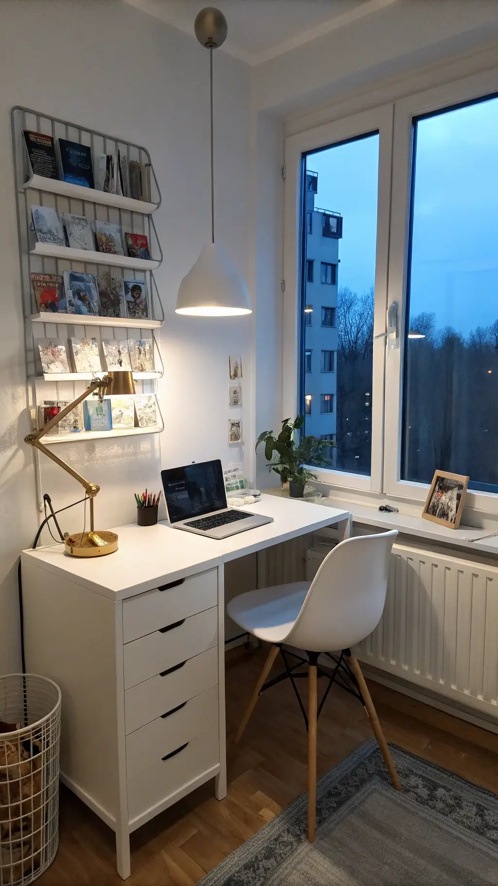 Scandinavian-inspired tiny office corner with white IKEA Alex desk, brass hardware, ghost chair, and magazine racks in powder-coated steel under a minimal pendulum lamp