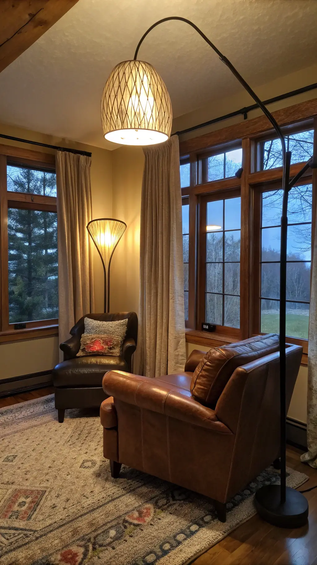 Warmly lit living room at dusk with layered lighting, brass reading lamp, LED accent walls, oversized pendant lights, and automated silk curtains on expansive windows.