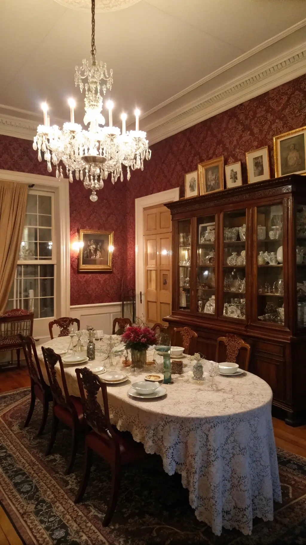 Dusk-lit formal dining room with chandelier glow over mahogany table, china cabinet teacups and silverware, family photos in gold frames on gallery wall, burgundy damask wallpaper