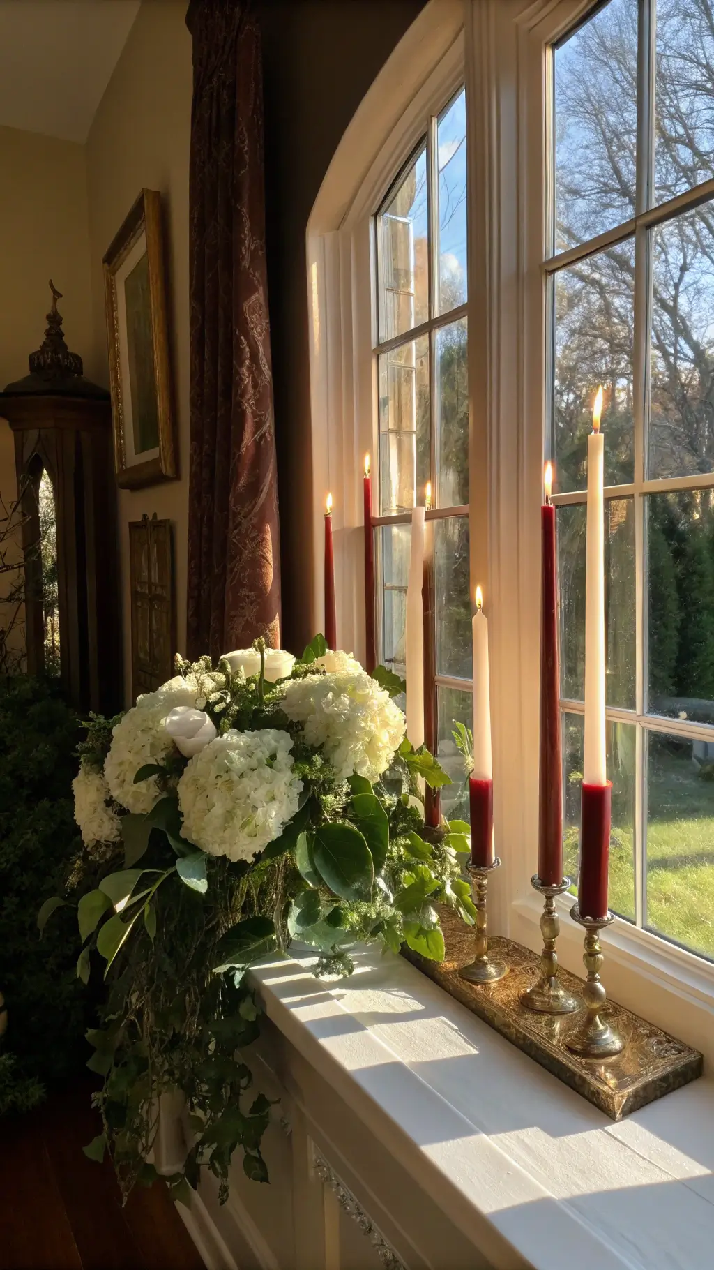Corner view of a traditional living room window alcove featuring a collection of Greek Orthodox Pascha candles on a vintage brass étagère, accented by white hydrangeas and trailing ivy under late afternoon sunlight.