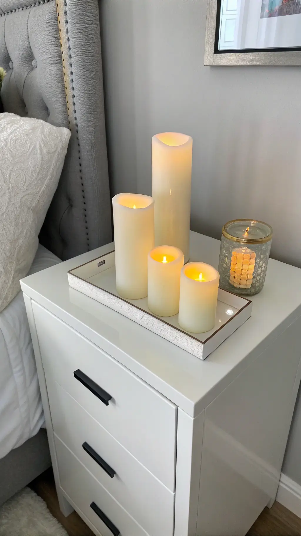 Modern minimalist bedroom with a trio of flameless LED Easter candles on clean-lined white lacquer nightstand, under dusk lighting against light gray walls.