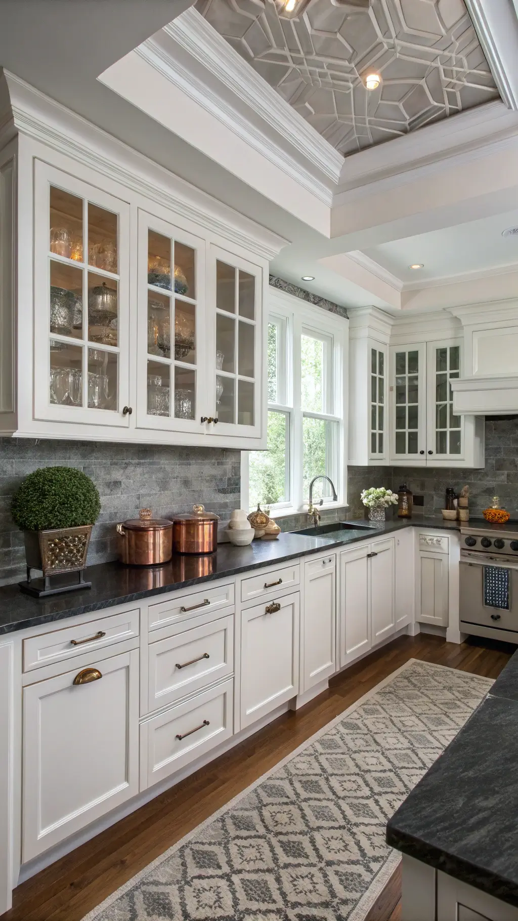 Transitional L-shaped kitchen with white inset cabinets, black leathered granite countertops, glass uppers, coffered ceiling, gray marble backsplash, copper cookware, and geometric runner.