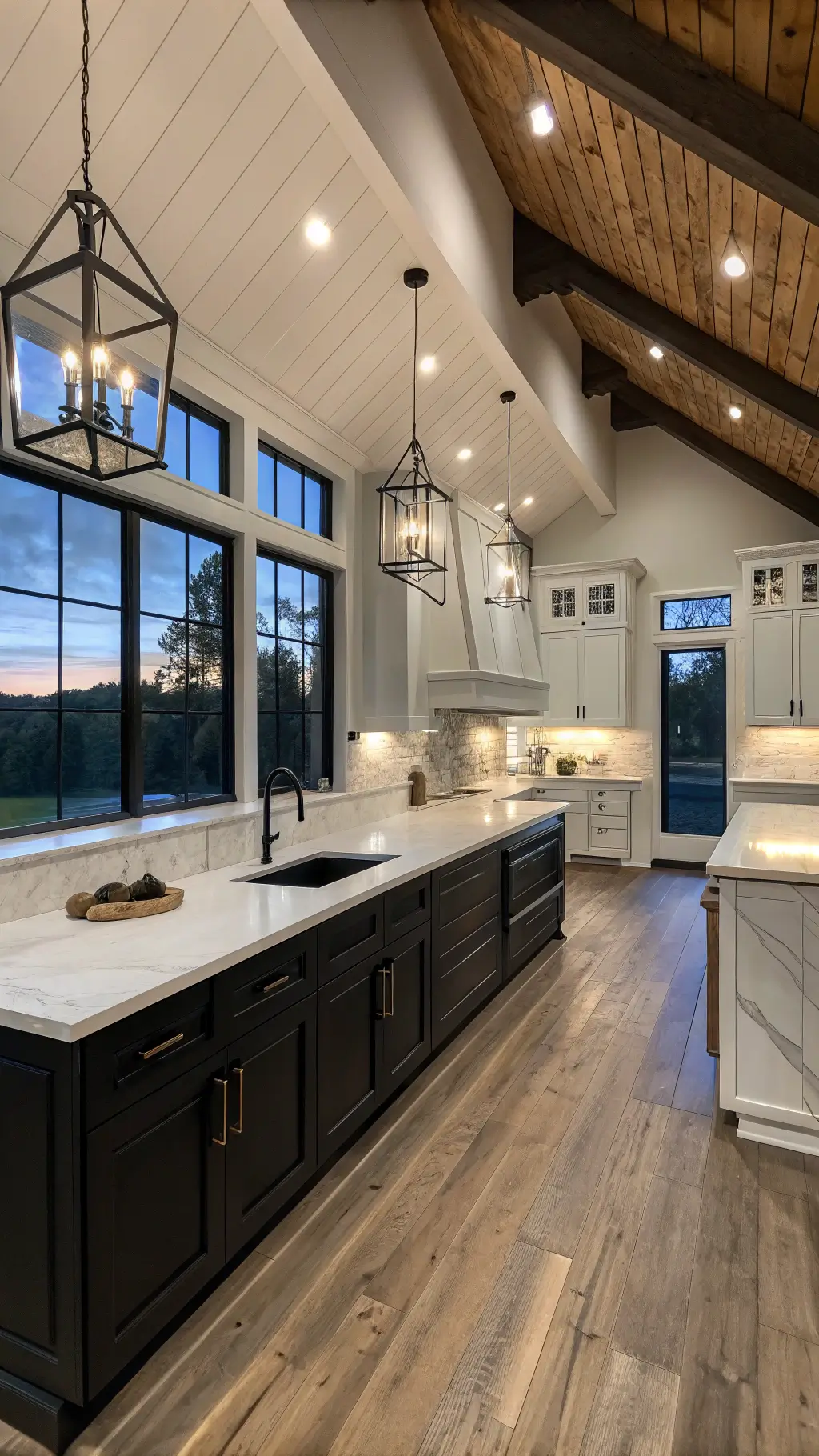 Contemporary farmhouse kitchen with 12ft trestled ceiling, two-tone black and white cabinets, industrial lighting, marble countertops, and rustic wood flooring at dusk.