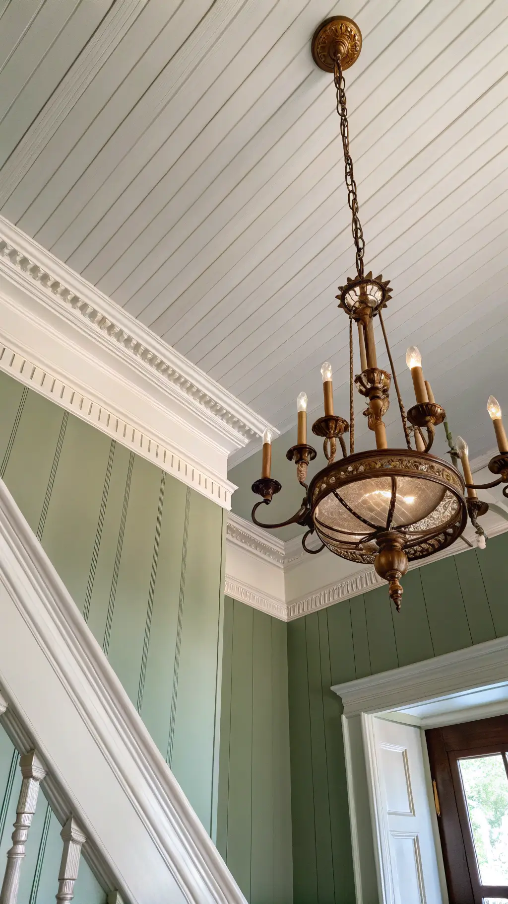 Low-angle view of ceiling and walls with white crown molding against sage green walls. A vintage brass chandelier hangs centrally, illuminated by afternoon light. Shiplap texture visible on the ceiling.