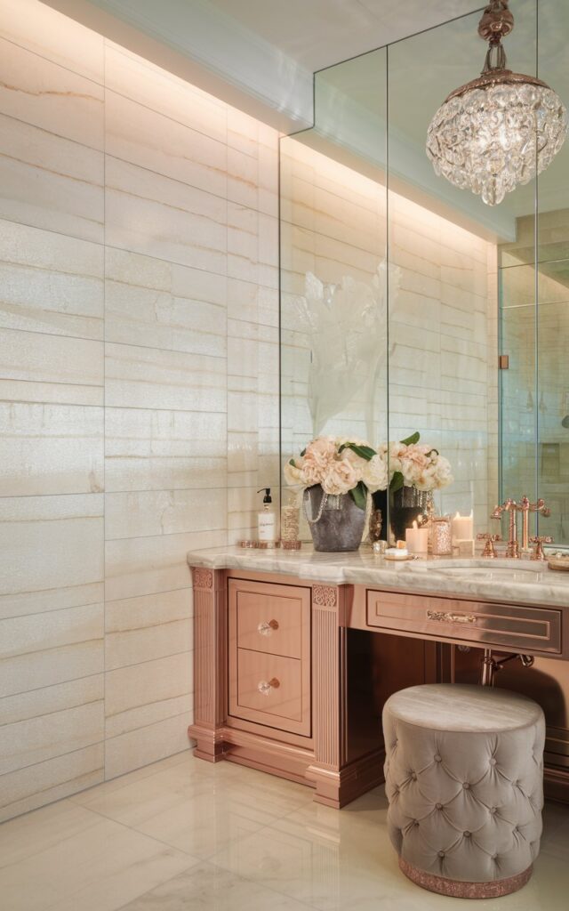 A photo of a luxurious bathroom with a glamorous vanity. The vanity has a marble countertop, rose-gold fixtures, and a cabinet. The wall behind the vanity is covered in large-format porcelain tiles with a subtle linen-textured pattern. The tiles are in soft ivory or warm greige tones and have a delicate sheen. A statement crystal pendant light hangs overhead. The room is illuminated by soft, ambient natural light. The space also features a velvet stool, scented candles, and a designer perfume bottle as accents.