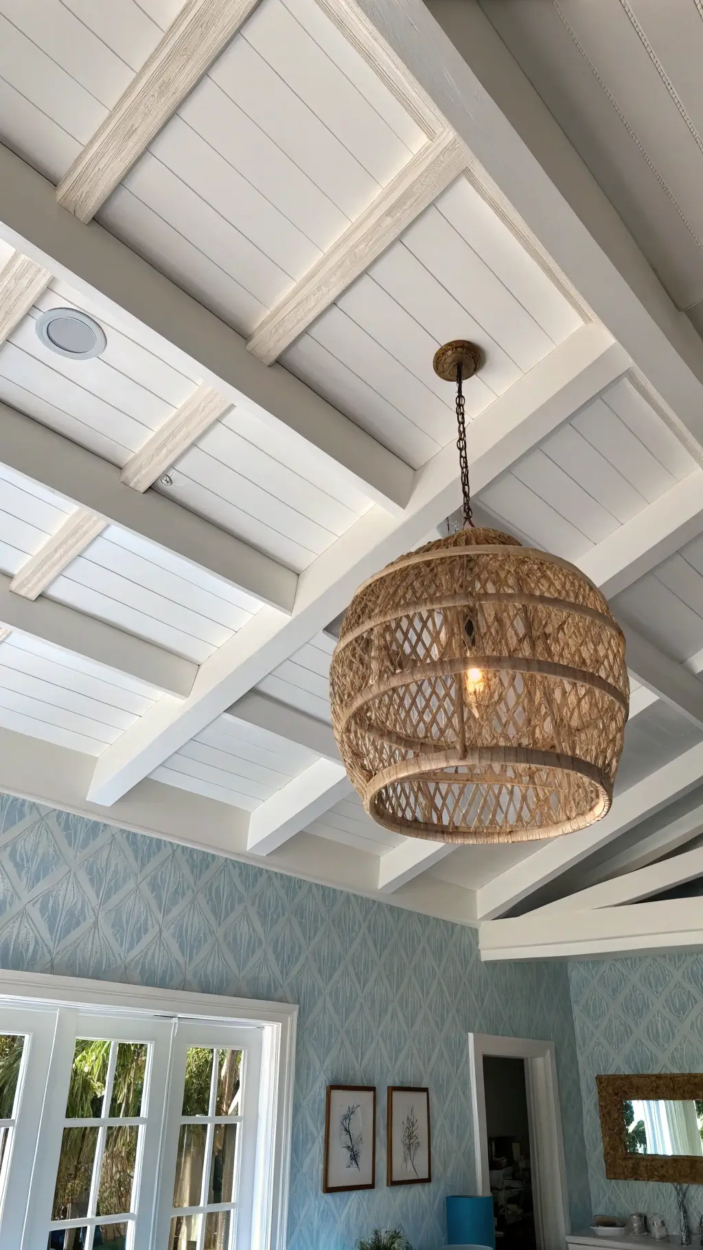 White wooden ceiling beams contrast with a light blue ceiling, as a large rattan pendant light casts intricate shadows. Upper walls with grasscloth wallpaper are partially visible, enhancing the architectural detail.