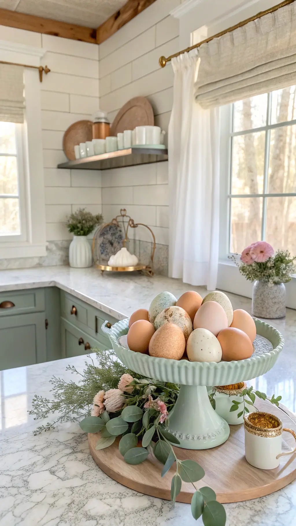 Bright farmhouse kitchen with shiplap walls, marble countertops, and morning light streaming through café curtains. Vintage enamel cake stand holds artisanal speckled eggs in sage, cream, and dusty blue. Fresh eucalyptus garland weaves through vintage teacups with pale pink ranunculus. Warm copper accents complement the airy composition.